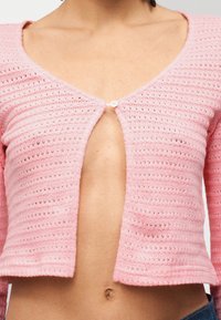 Γυναίκα που φοράει ένα ροζ πλεκτό κοντό ζακετάκι με ένα κουμπί κλειστό, αφήνοντας ακάλυπτη τη μέση και τον ομφαλό, μπροστά σε απλό φόντο.