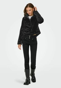 Femme portant une veste en fausse fourrure noire, un jean skinny noir taille haute et des bottes noires mi-mollet avec des détails à boucle, posant avec la main dans les cheveux.