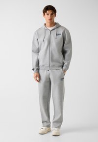Sweat à capuche gris zippé et pantalon de survêtement assorti. Matière douce et texturée. Orné d'un logo bleu sur la poitrine et la jambe du pantalon. Design décontracté.