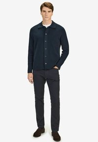 Navy knit cardigan with button closure, designed with a spread collar and long sleeves, paired with dark trousers and brown suede shoes.