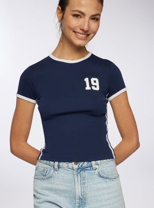 Giovane donna sorridente, indossa una maglietta aderente blu navy con bordi bianchi e il numero 19 sul petto, abbinata a jeans in denim azzurro chiaro.