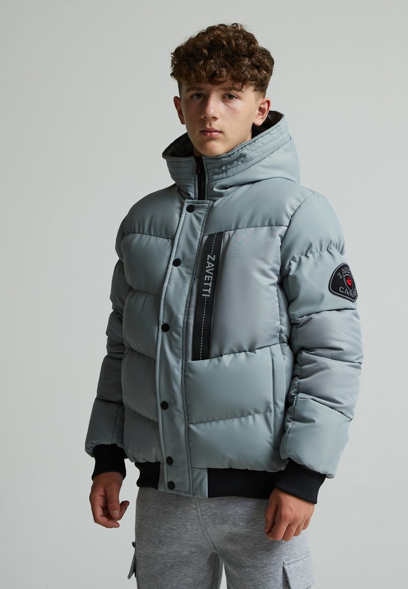 Veste grise matelassée avec col haut et accents noirs. Dotée d'une fermeture éclair, de grandes poches avant et d'un patch logo sur la manche.