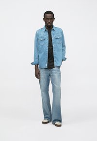 Světle modrá džínová košile s předními kapsami, zapínáním na knoflíky, nošená přes černé tričko, kombinovaná s rozšířenými světle modrými džínami a béžovými botami.