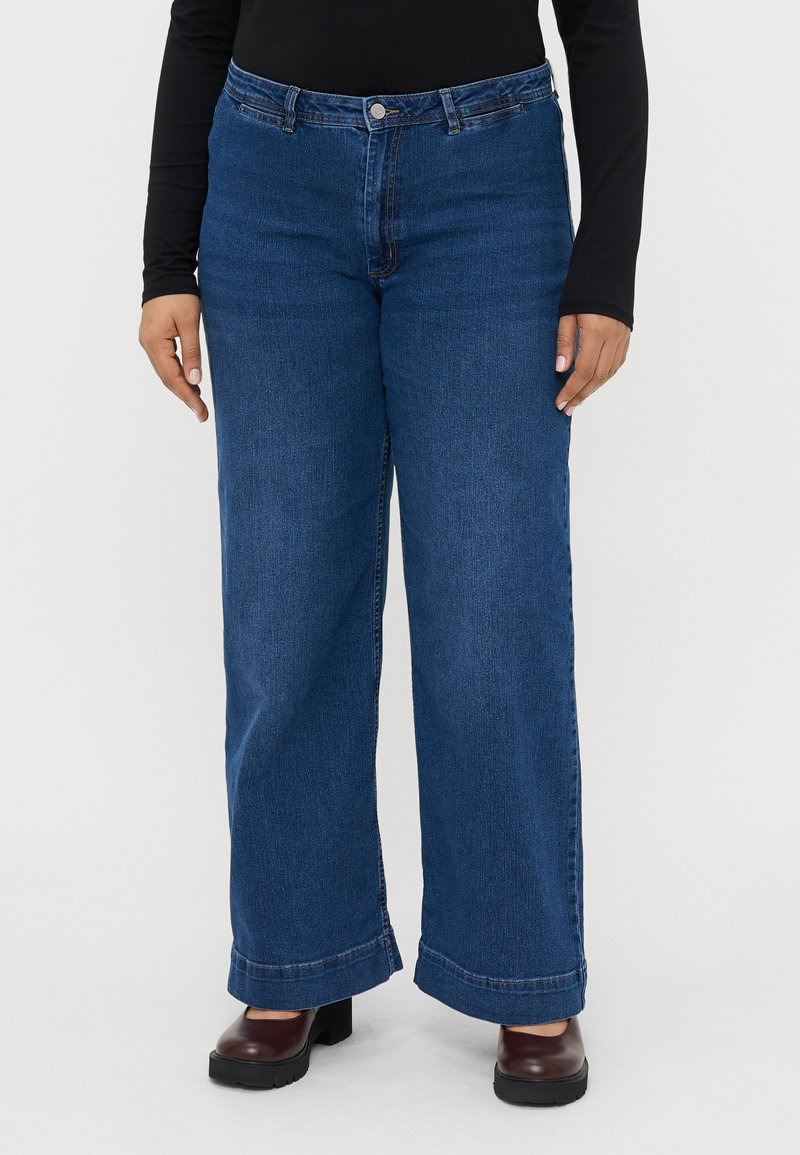 Jeans de perna larga em denim azul escuro, com fecho de botão, design clássico de cinco bolsos e bainhas viradas para cima. Combinados com sapatos pretos.