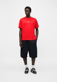 Άνδρας που φοράει κόκκινο κοντομάνικο μπλουζάκι, μαύρα σορτς μέχρι το γόνατο, λευκές κάλτσες και μαύρα αθλητικά παπούτσια Nike, στέκεται μπροστά σε απλό λευκό φόντο.