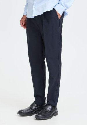 Man draagt zwarte nette schoenen en donkerblauwe formele broek met handen in de zakken, vanaf de taille geknipt, staand op een witte achtergrond.