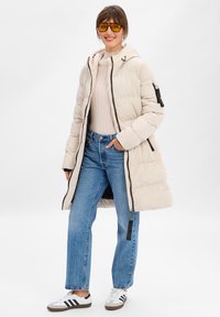 Jasno beżowy, pikowany długi płaszcz z kapturem, czarnym zamkiem błyskawicznym i bocznymi kieszeniami. Noszony na jasnym swetrze z prążkami i niebieskich dżinsach, zestawiony z białymi trampkami.