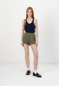 Námornícky top bez rukávov s bielym lemom, olivovo zelené šortky, čierne mokasíny a svetlo šedé ponožky. Model drží hnedú kabelku cez rameno.