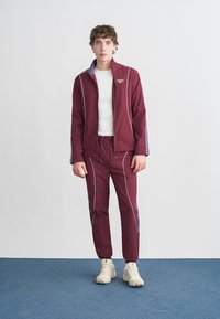 Bordó színű futódzseki fehér csíkozással és lila részletekkel, hozzáillő nadrággal párosítva. A modell fehér inget és világos színű sportcipőt visel.