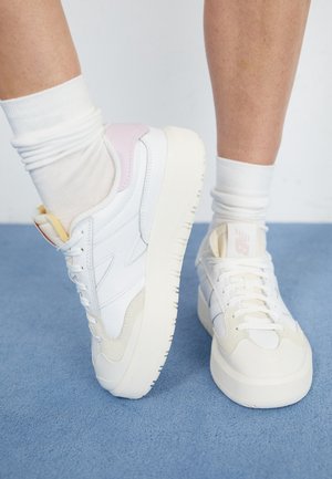 Baskets à plateforme blanches avec accents beige et rose portées avec des chaussettes blanches sur un tapis bleu, montrant les vues de face et de côté des pieds.