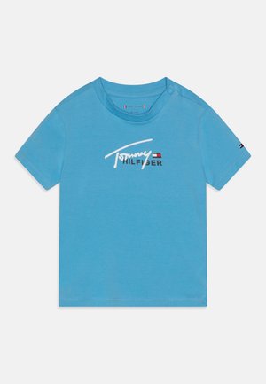 T-shirt bleu à manches courtes pour tout-petit avec le logo Tommy Hilfiger et un drapeau sur la poitrine ainsi qu'un petit drapeau sur la manche gauche. Deux boutons-pression à l'épaule.