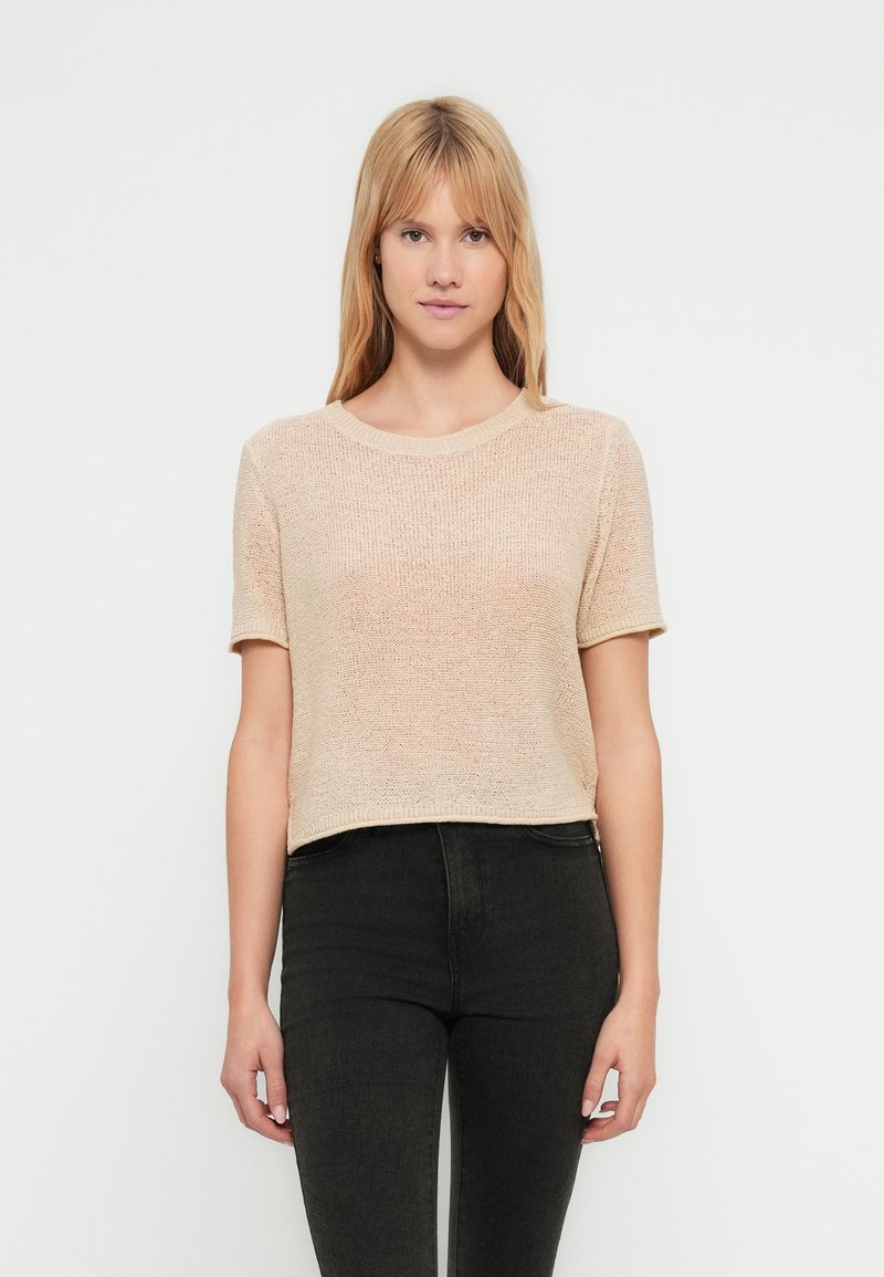 T-shirt cropped em malha bege claro com mangas curtas, decote redondo e textura subtil. Combinada com jeans escuros.