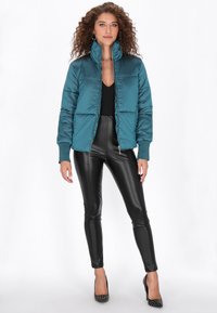 Turkusowa puchowa kurtka z wysokim kołnierzem i zapięciem na zamek, w połączeniu z czarnymi błyszczącymi legginsami. Teksturowany materiał z pikowanym wzorem i subtelnym blaskiem.