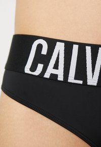 Svart bikinitruse med en bred elastisk midjebånd med "CALVIN" i hvit skrift. Glatt, elastisk stoff med en tettsittende design.
