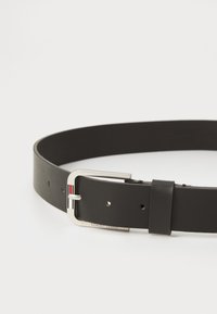 Ceinture en cuir noir avec une boucle en argent, ornée d'une petite touche rouge et blanche. Sangle texturée avec plusieurs trous d'ajustement.