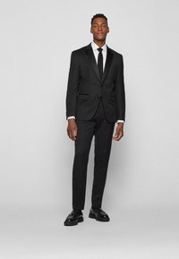 Completo nero con rever in raso, design slim-fit, pantaloni formati e camicia bianca. La cravatta nera e le scarpe nere lucide completano l'outfit.