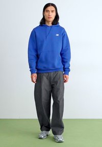 Blå hettegenser med frontlomme og logo, sammen med løstsittende grå bukser. Modellen har også på seg sølvfargede joggesko på et grønt gulv.