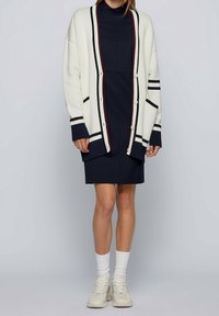 Bílý kardigan s námořnickými a burgundskými pruhy, s volným střihem, knoflíkovými uzávěry a žebrovanými manžetami, nošený přes námořnické šaty a bílé ponožky.