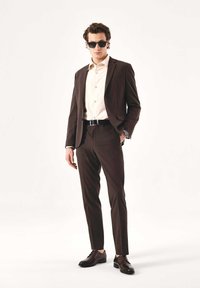 Abito marrone scuro con pantaloni a taglio slim e giacca abbinata. Camicia di colore chiaro e scarpe nere, completati da occhiali da sole scuri.