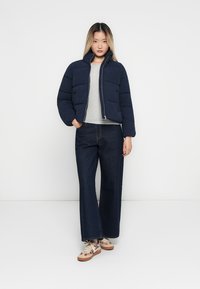 Marineblå jakke med hette og glidelås, med høy krage. Brukt over en lys grå skjorte og kombinert med vide mørke jeans og mønstrede sneakers.