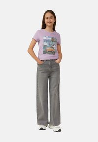Lila T-Shirt mit einem bunten Grafikdesign, kombiniert mit locker sitzenden grauen Jeans und weißen Sneakers. Einfaches, lässiges Outfit.
