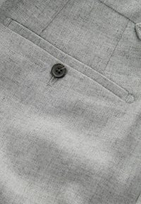 Tela gris con un primer plano de un bolsillo con un botón oscuro cosido. La textura parece suave con un patrón sutil.