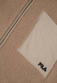 Beige fleece stof met een gestructureerde afwerking, gecombineerd met een glad paneel in beige met een zwart "FILA" logo in de hoek.
