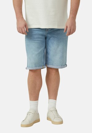 Man draagt lichtblauwe opgerolde denim shorts, witte crew sokken en witte sneakers, staand tegen een effen achtergrond.