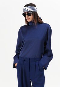 Top blu navy con collo alto e maniche lunghe, abbinato a pantaloni a pieghe coordinati. Accessoriato con una fascia a righe e occhiali da sole neri.