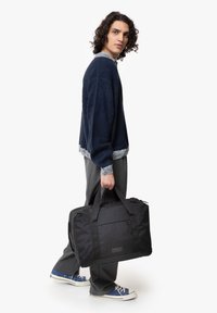 Sac de sport en nylon noir avec poignées supérieures et poche avant, présentant un tissu texturé et une forme rectangulaire, porté par une personne.