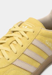 Κίτρινο σουέντ αθλητικό παπούτσι με μπεζ δερμάτινες ρίγες, κορδόνια με υφή και ετικέτα με λογότυπο. Διαθέτει λάστιχο στη σόλα και ραμμένες λεπτομέρειες.