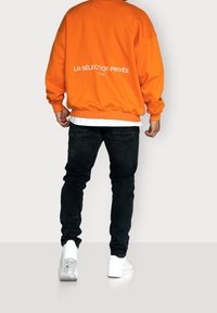 Felpa arancione oversize con testo bianco sul retro, abbinata a jeans neri e sneakers bianche. Il tessuto appare morbido con una finitura texture.