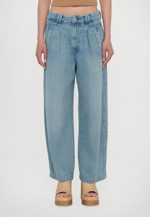 Femme portant un jean bleu clair, taille haute et ample, et des sandales beige à plateforme ouvertes sur les orteils, debout devant un fond blanc uni.