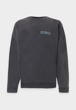 Model trägt einen schwarzen übergroßen Pullover und weit geschnittene blaue Jeans, steht mit den Händen in den Taschen vor einem schlichten Hintergrund.