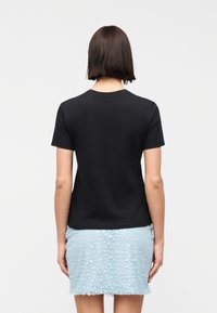 Γυναίκα με καστανά κοντά μαλλιά που φοράει απλό μαύρο κοντομάνικο μπλουζάκι και ανοιχτό μπλε υ φασμάτινη φούστα, στέκεται με την πλάτη προς το μέρος μας μπροστά σε λευκό φόντο.