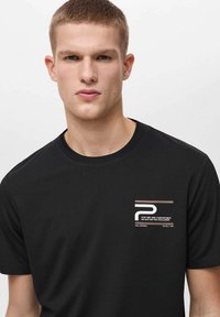 T-shirt nera a maniche corte con tessuto texturizzato, colletto rotondo e stampa grafica sul lato sinistro del petto in tonalità di bianco e marrone.