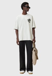 Hombre con gafas de sol negras, camiseta blanca de gran tamaño con estampado, pantalones negros, zapatillas blancas, sosteniendo una bolsa de lona beige al costado.