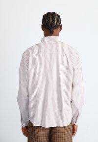 Person mit geflochtenem Haar, der ein weißes Hemd mit feinen roten Streifen und braunen Karohosen trägt, steht mit dem Rücken zu einem schlichten weißen Hintergrund.