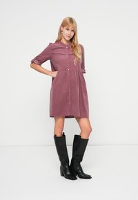 Sukienka zapinana na guziki z tkaniny w kolorze mallowym, z krótkimi podwiniętymi rękawami, pełną spódnicą i delikatną lamówką na wysokości klatki piersiowej. Czarne kozaki do kolan.