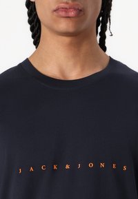 Marineblå bomulds T-shirt med rund halsudskæring og korte ærmer, med "JACK & JONES" logo i orange på tværs af brystet. Glat tekstur.