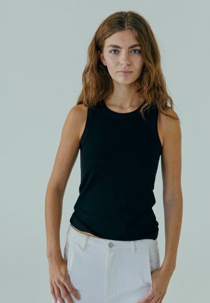 Haut sans manches noir avec un col rond, fabriqué en tissu doux. Le mannequin l'associe avec un pantalon blanc, affichant une silhouette ajustée.