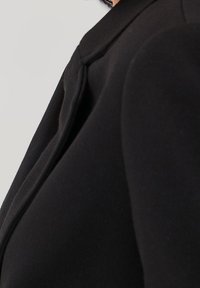 Musta bleiseri, jossa on muotoiltu fit, näyttävä kaulus ja teksturoitu kangas. Yksityiskohtiin kuuluvat siistit saumat ja huomaamaton ompelu.
