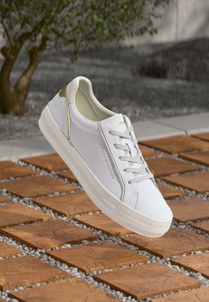 Adidas alb cu accente aurii plutind deasupra unei podele din gresie și piatră în aer liber, cu un copac în fundal.