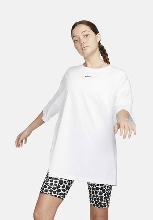 Joven con el pelo trenzado lleva una camiseta blanca Nike de gran tamaño y pantalones cortos de ciclismo negros y blancos con motas sobre un fondo liso.