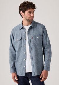 Blue patterned shirt made of textured fabric, featuring a button-front, two chest pockets, and a pointed collar, worn over a white shirt.
