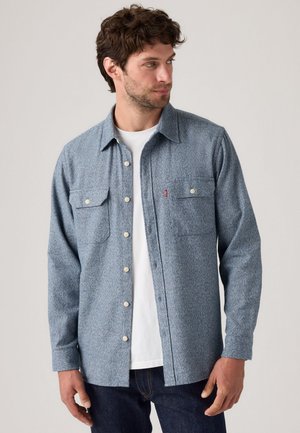 Blue patterned shirt made of textured fabric, featuring a button-front, two chest pockets, and a pointed collar, worn over a white shirt.