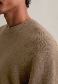 Pull en tricot marron texturé avec un col rond et des détails côtelés sur les épaules, mettant en avant un tissu doux et chaud.