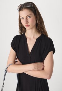 Fekete V nyakú ruha rövid ujjakkal, könnyű anyagból, laza szabású, texturált részletekkel. A modellen napszemüveg és kék pántos táska van.