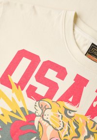 Camiseta de color crema con letras "OSAK" en neón rosa y un gráfico de tigre colorido con acentos de rayos. Textura de algodón suave.