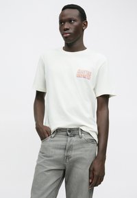 Νεαρός άνδρας με σκούρο δέρμα και κοντά μαλλιά, φορώντας λευκό t-shirt με το θέμα του "David Bowie" και γκρι τζιν, στέκεται με το ένα χέρι στην τσέπη του, μπροστά σε λευκό φόντο.